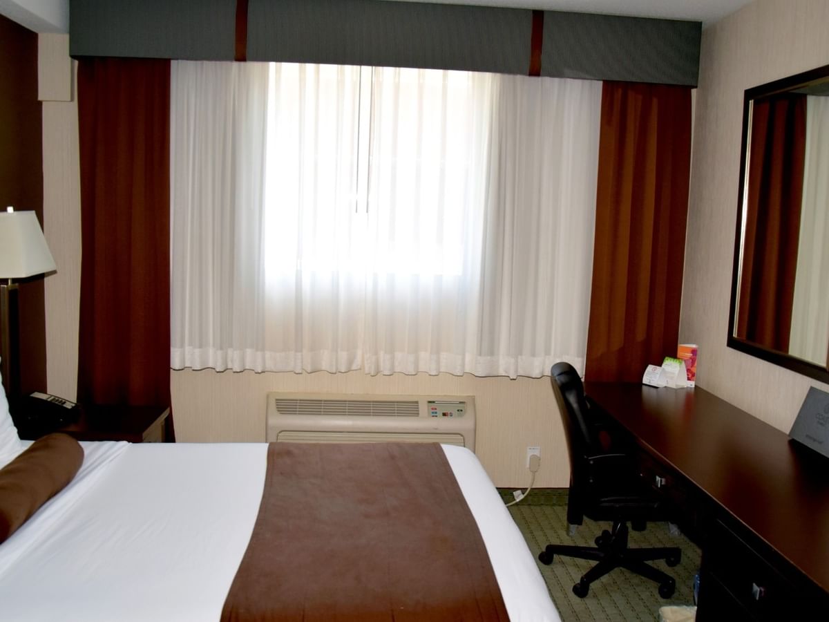 Hotel room with bed, desk, chair, lamp, TV, and window with curtains.