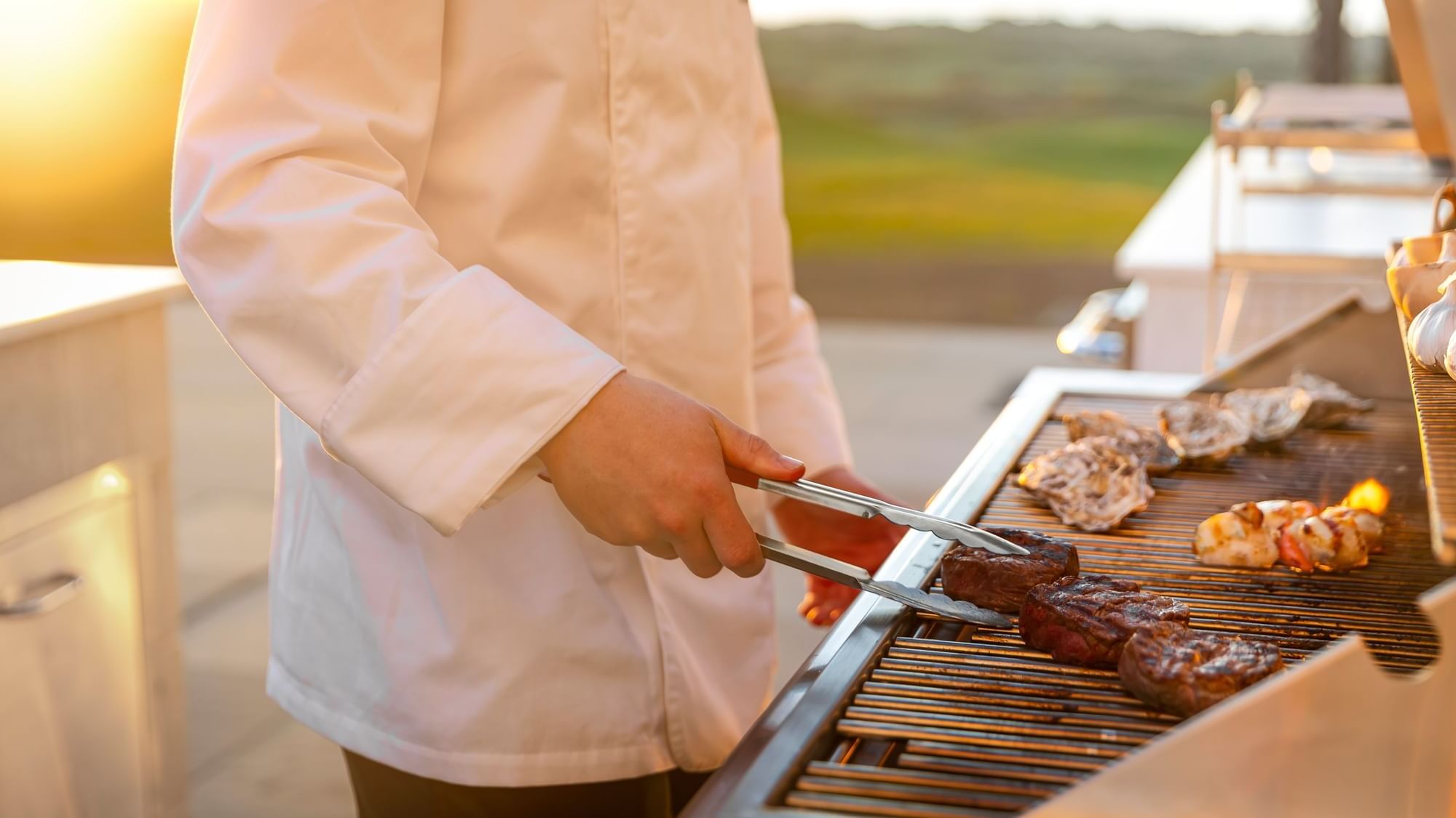 A chef with tongs grilling steaks, oysters, and scallops on a barbecue at Dunluce Lodge, outdoors