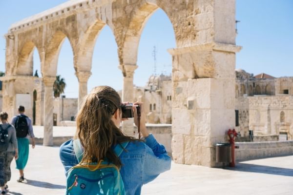 A woman in a denim jacket and backpack has her back to the camera as she takes a photo of old stone arches. 