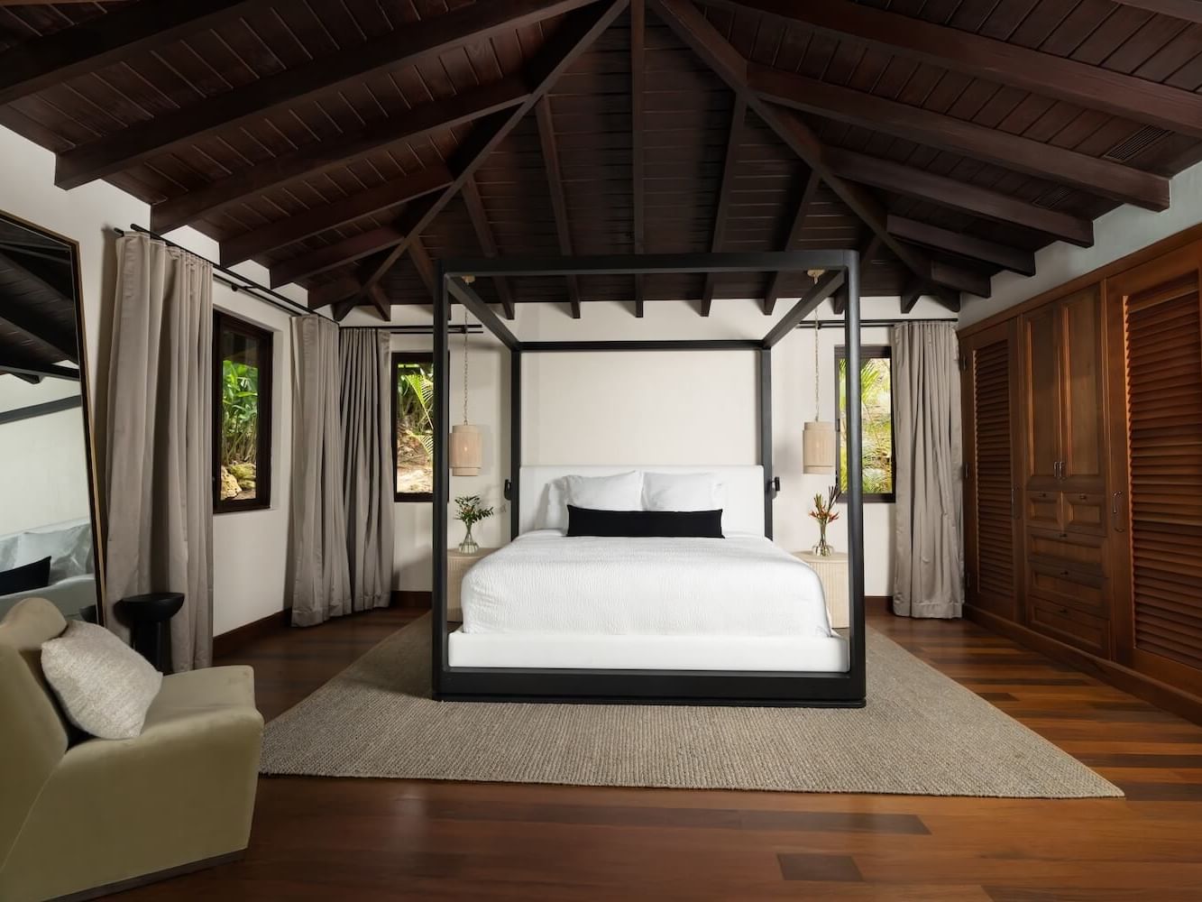 Four-poster king bed with white linens and rattan pendant lights, framed by windows with tropical greenery and a wood wardrobe. Warm, low light falls under a beamed ceiling.