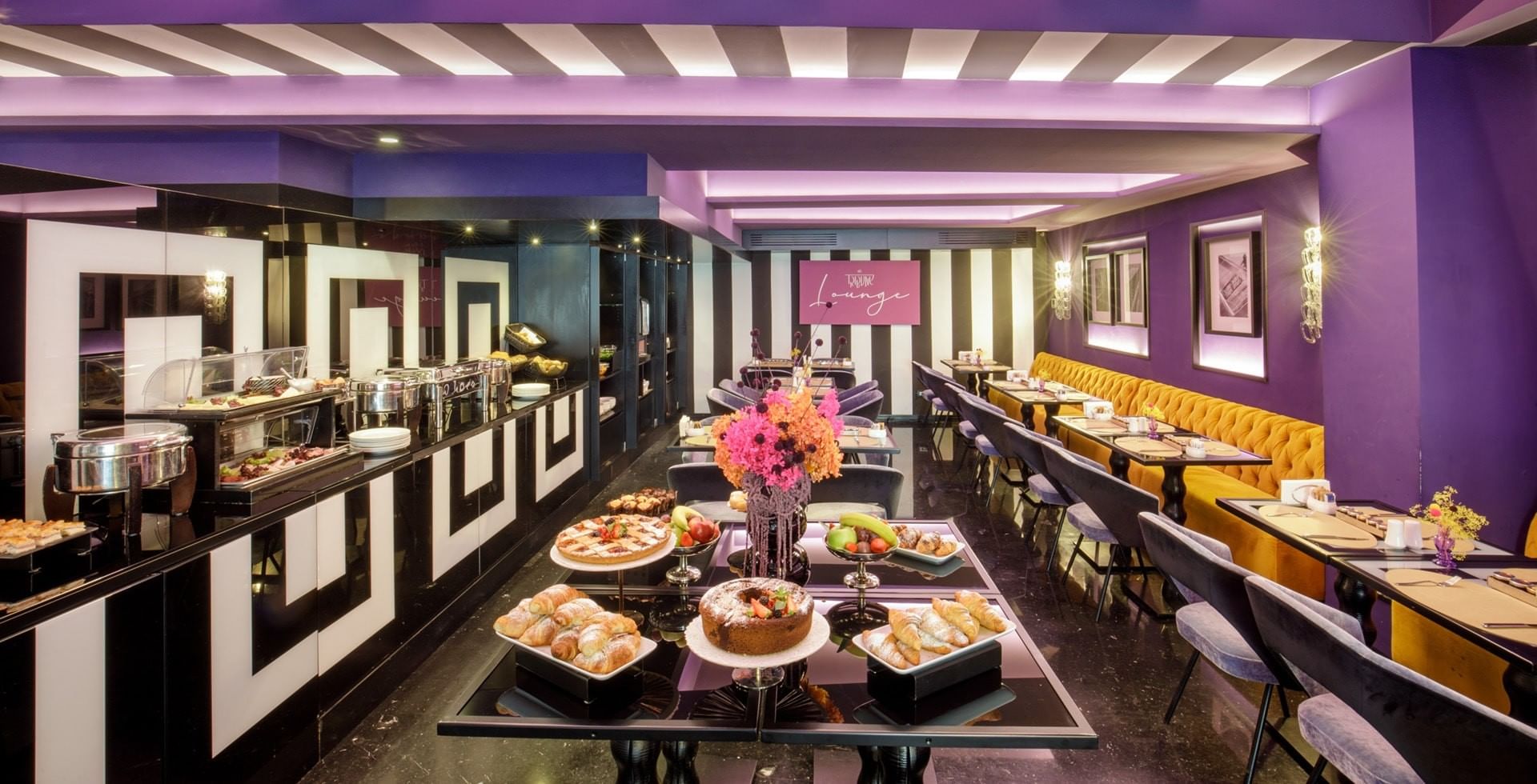 Elegant dining lounge at Tribune Hotel Rome with vibrant décor, velvet seating, and buffet breakfast offering fresh pastries, fruit, and cakes.
