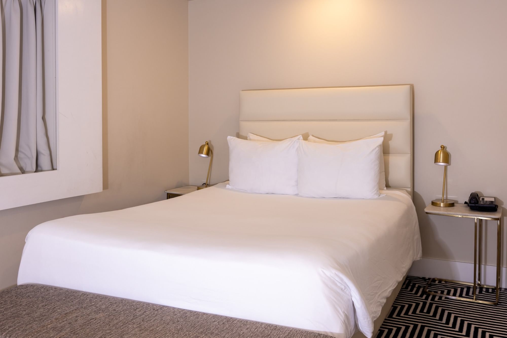 A neatly made bed with white linens and pillows in a room with beige walls and a geometric patterned floor.