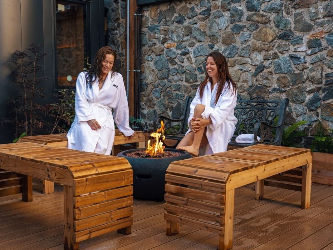 Two women in white robes relax on wooden loungers beside a fire pit on a stone-walled outdoor terrace.