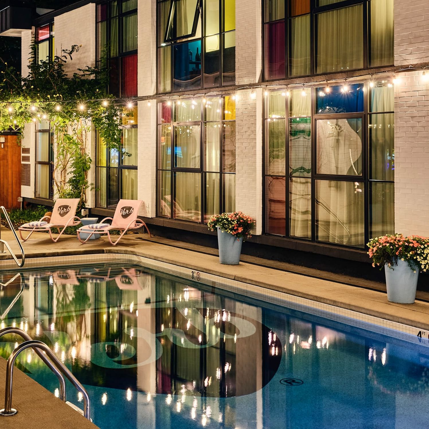 Modern hotel pool illuminated at dusk with colorful glass windows and lounge chairs reflecting in the water.