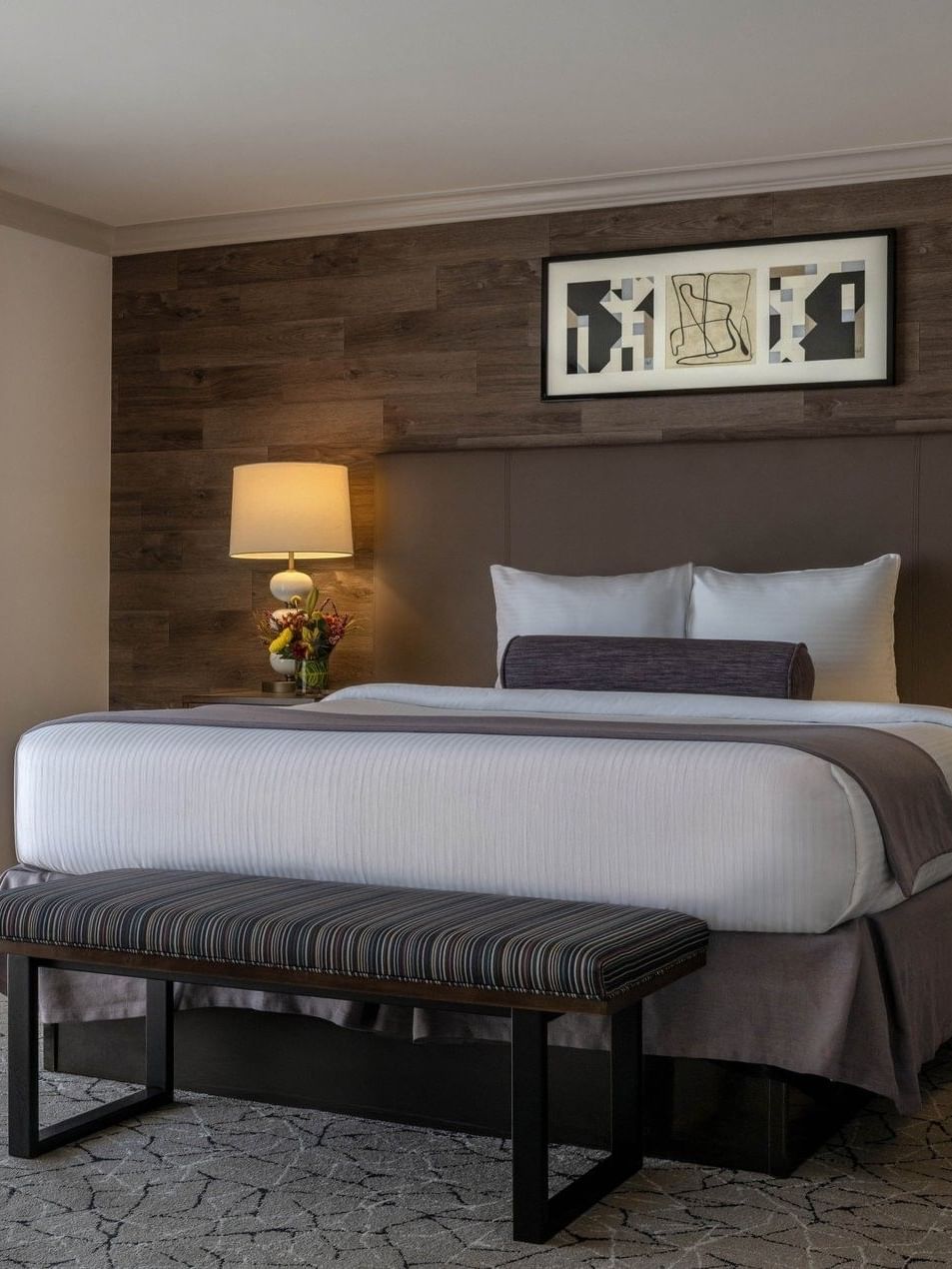 Striped bench by a plush bed under a wood-paneled wall with framed art in Deluxe Room at Warwick Denver