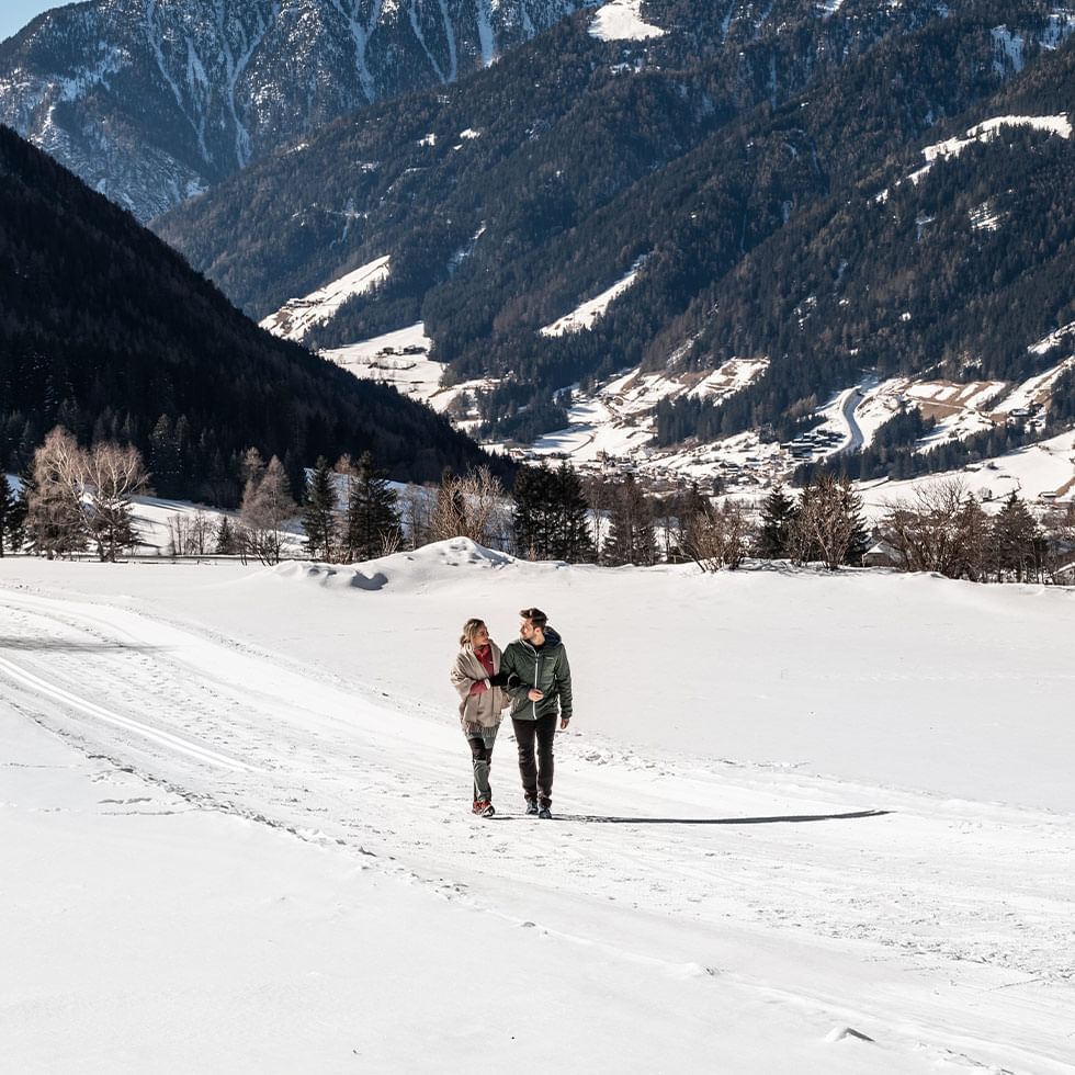 Due persone camminano su un sentiero innevato con montagne innevate sullo sfondo.