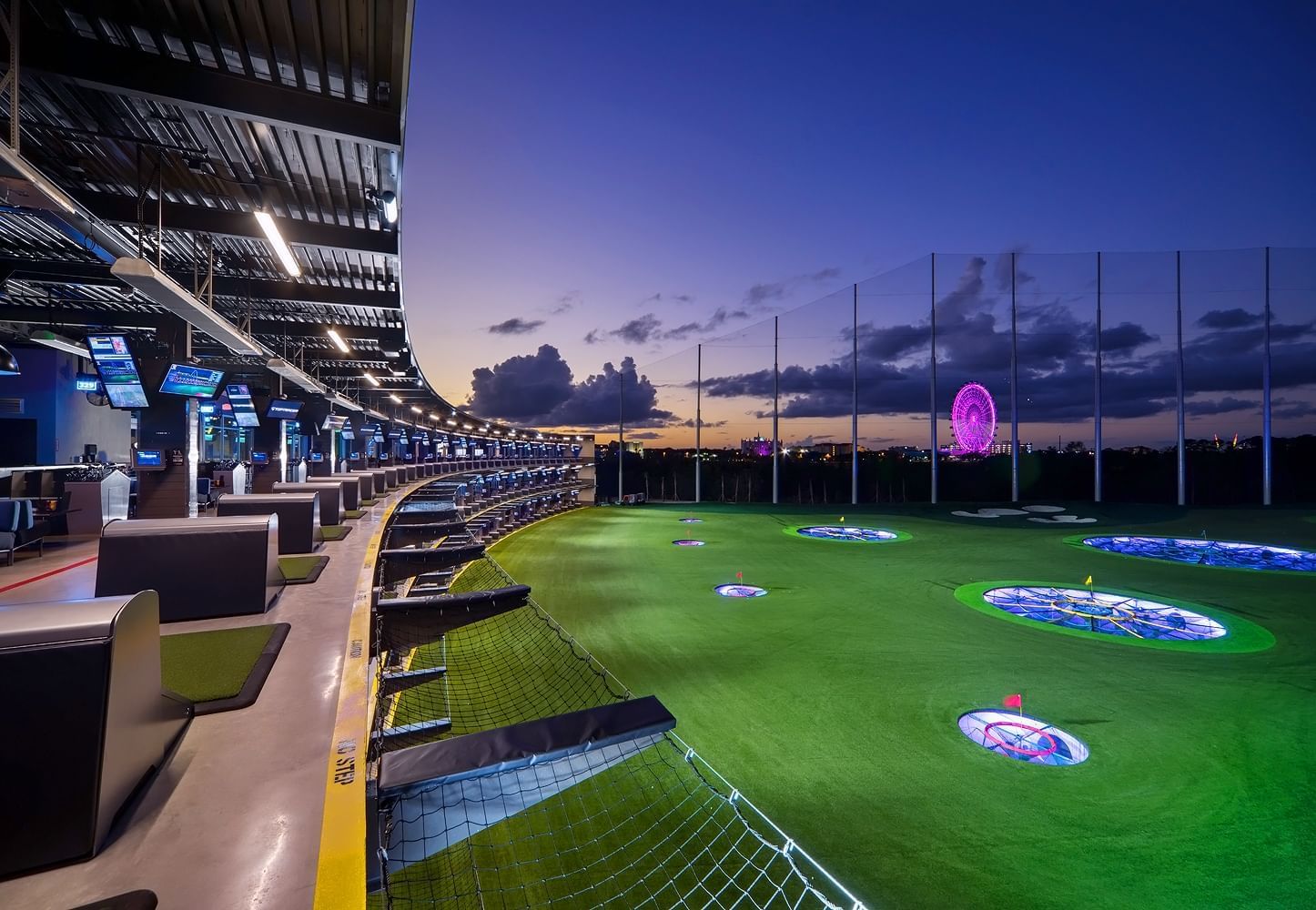 A large driving range at twilight with a long curving covered patio area with individual bays.