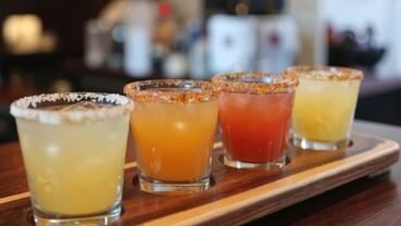 Four colorful sips marg glasses on a wooden tray.