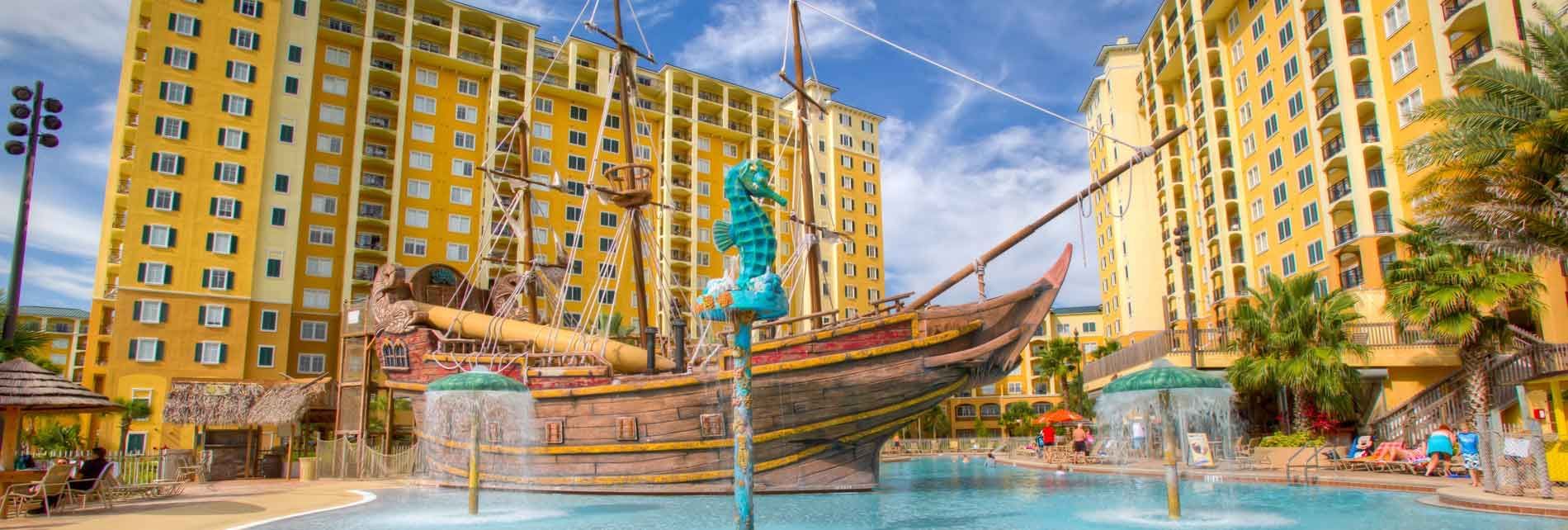 A large pool with fountains and a pirate ship in front of tall yellow buildings.