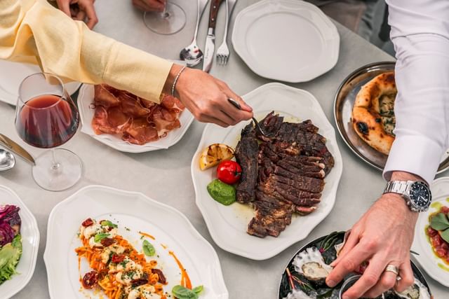 Close-up food view at Elysium Manly with hands by a steak platter surrounding pasta and oysters under bright light
