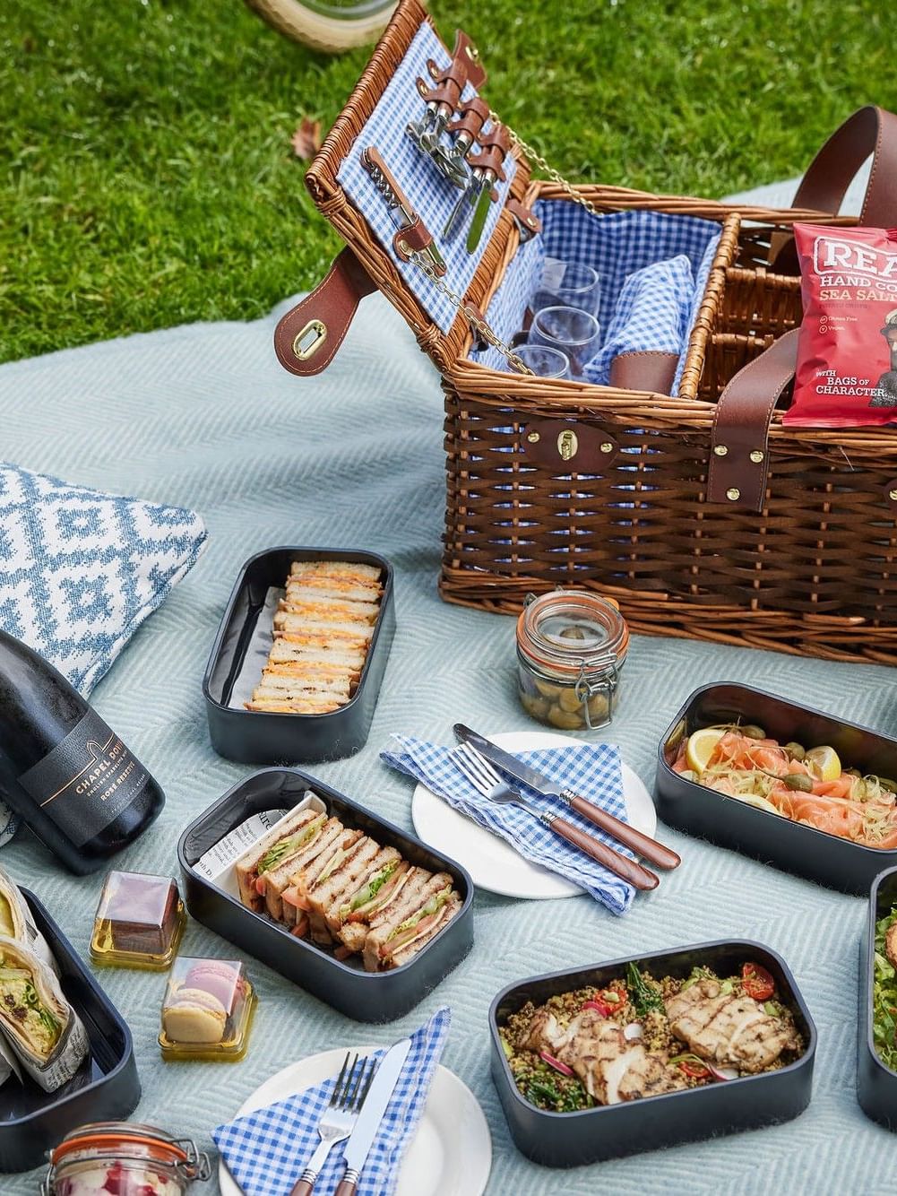 Picnic setup with wicker basket, sandwiches, fruit, crackers, and wine bottle on a blanket on grass.