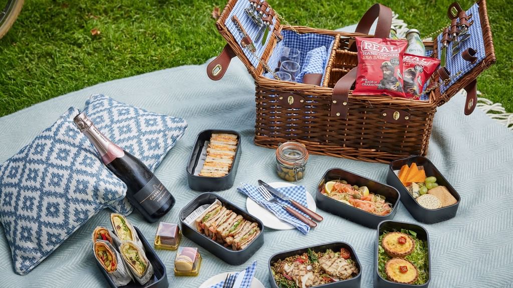 Picnic setup with wicker basket, sandwiches, fruit, crackers, and wine bottle on a blanket on grass.