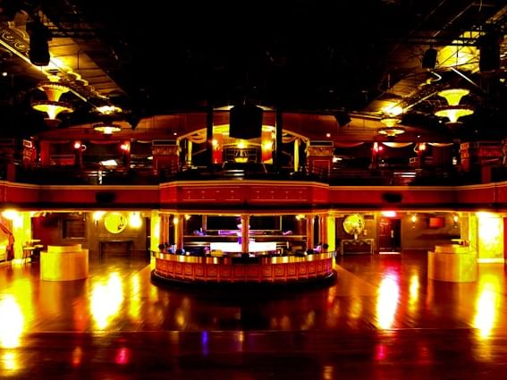 A dimly lit dance hall with a large dance floor, a central bar, and a balcony with red curtains.