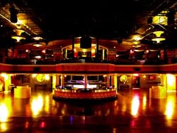 A dimly lit dance hall with a large dance floor, a central bar, and a balcony with red curtains.