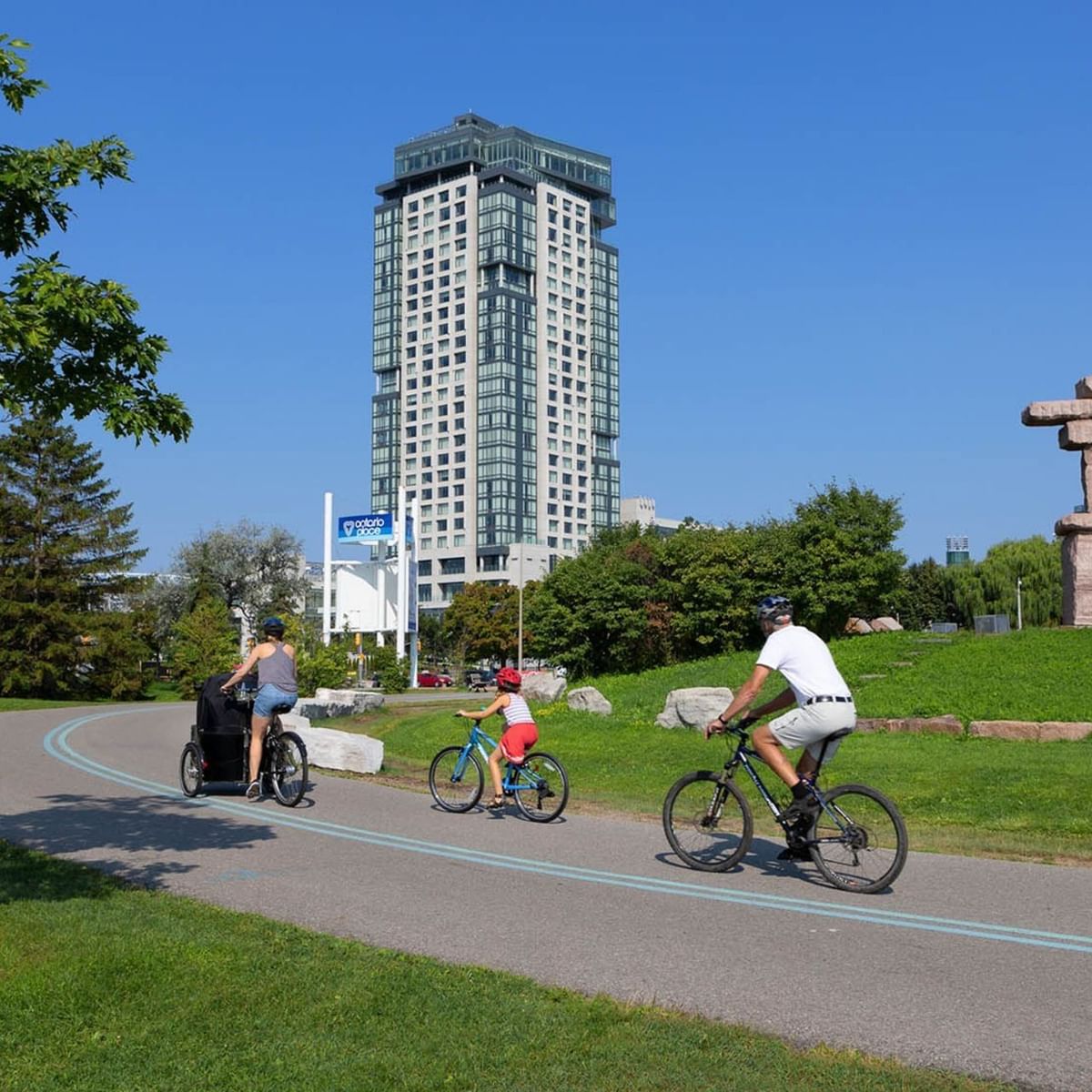 Three cyclists riding on a paved path through a green park with Hotel X Toronto in the background