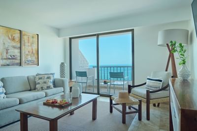 Living room with ocean view through glass doors, featuring couch, coffee table, chairs, and artwork.