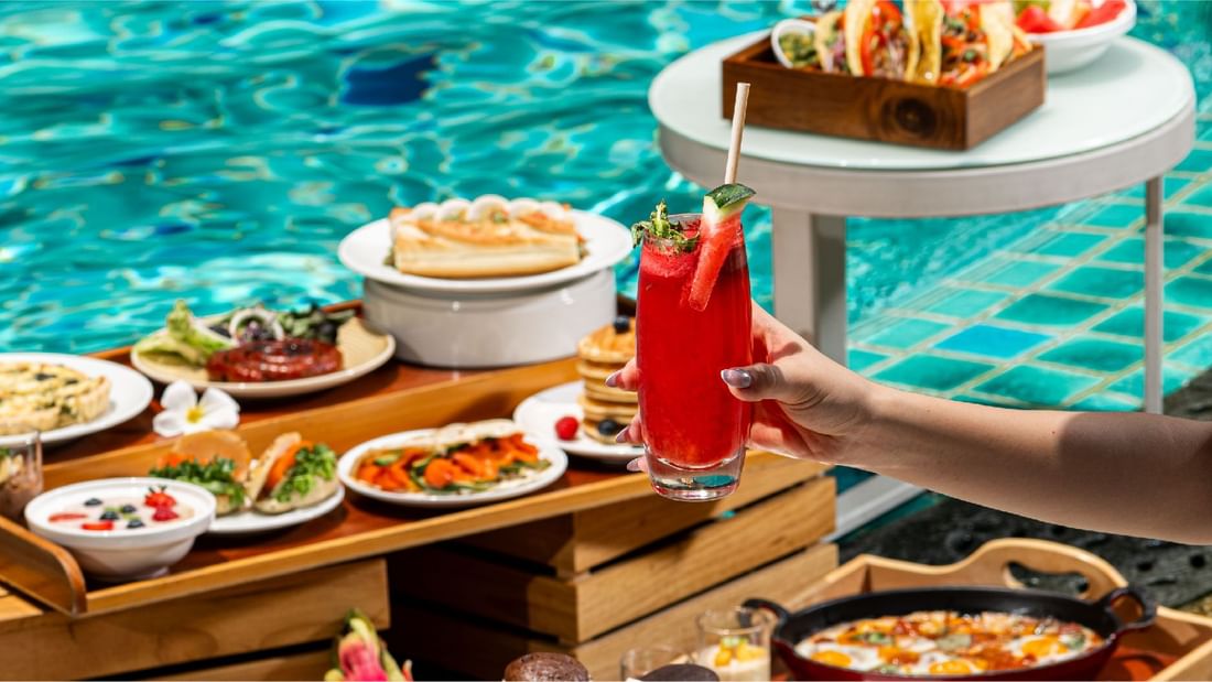 A hand holding a refreshing drink by a poolside brunch spread, offering delightful dining at Sunway Hotel Pyramid