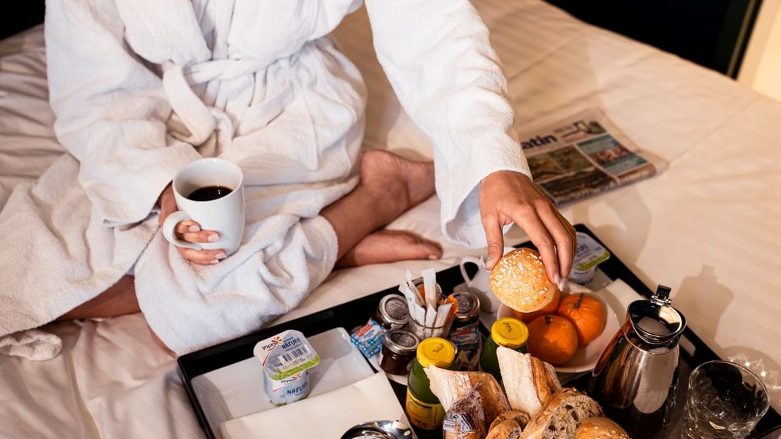 Femme en peignoir sur un lit avec un plateau de petit-déjeuner et une tasse de café.