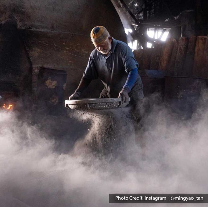 Worker sieving charcoal dust in My Charcoal Factory, surrounded by a cloud of dust in the dim light - Lexis Suites Penang