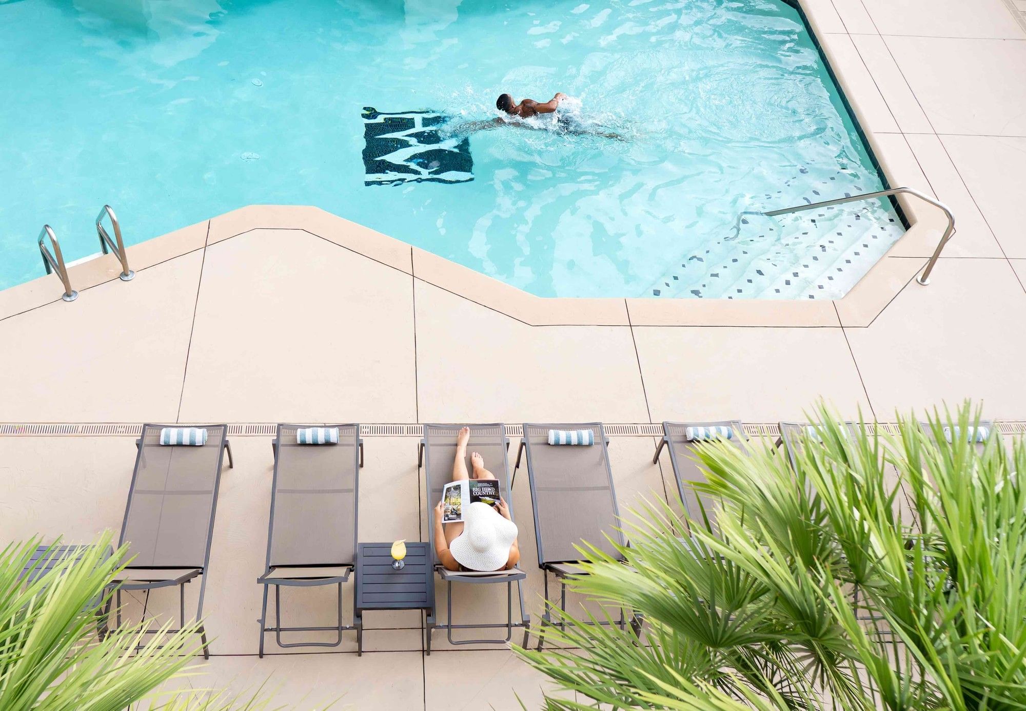 Aerial view of a pool with a person swimming and another person relaxing on lounge chairs.