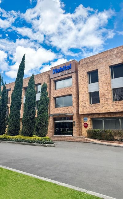 Exterior view of Habitel Prime hotel with brick facade, tall green trees, and a clear blue sky with scattered clouds