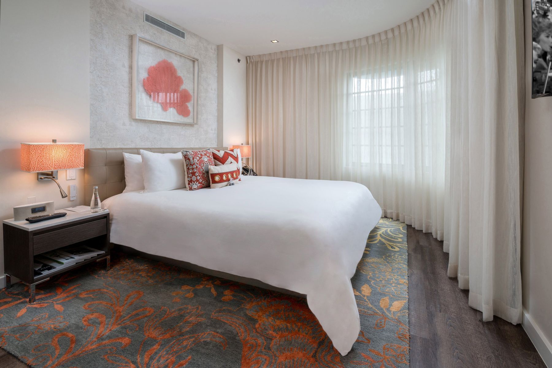 A wide-angle view of a hotel room with a king-sized bed, nightstand, and artwork on the wall.