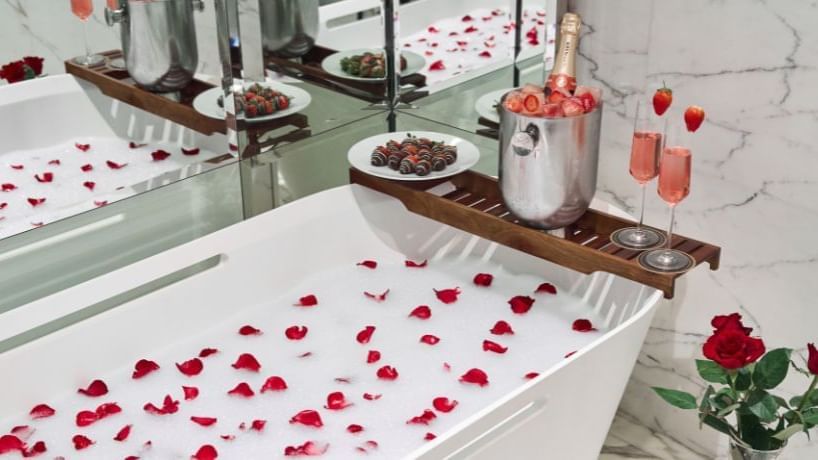 Luxurious bathtub with rose petals, a wooden tray holding champagne, and strawberries in a bathroom at Paramount Hotels