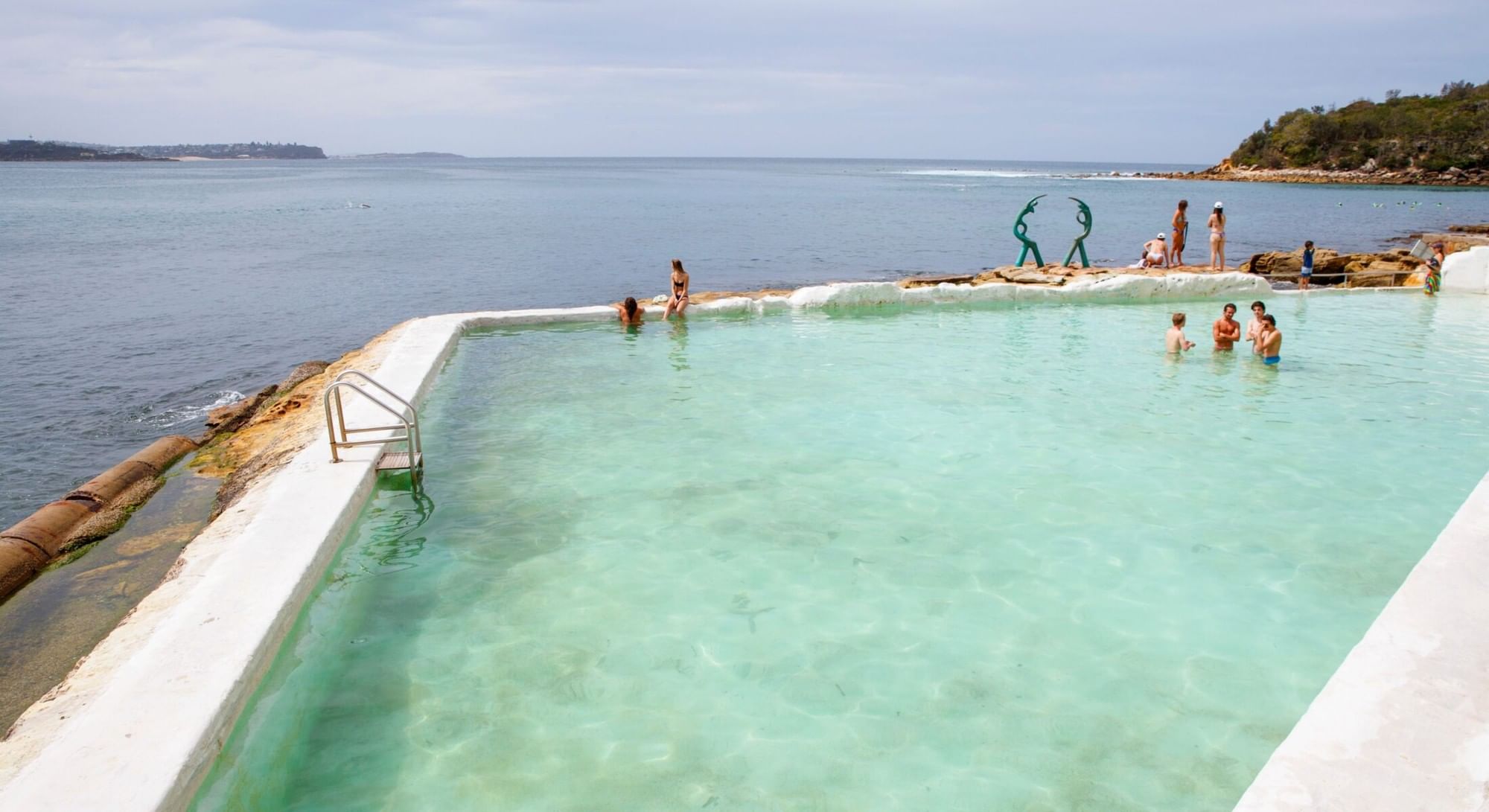 Fairy Bower Pool at Elysium Hotels is between a concrete walkway and the ocean with swimmers and a green sculpture