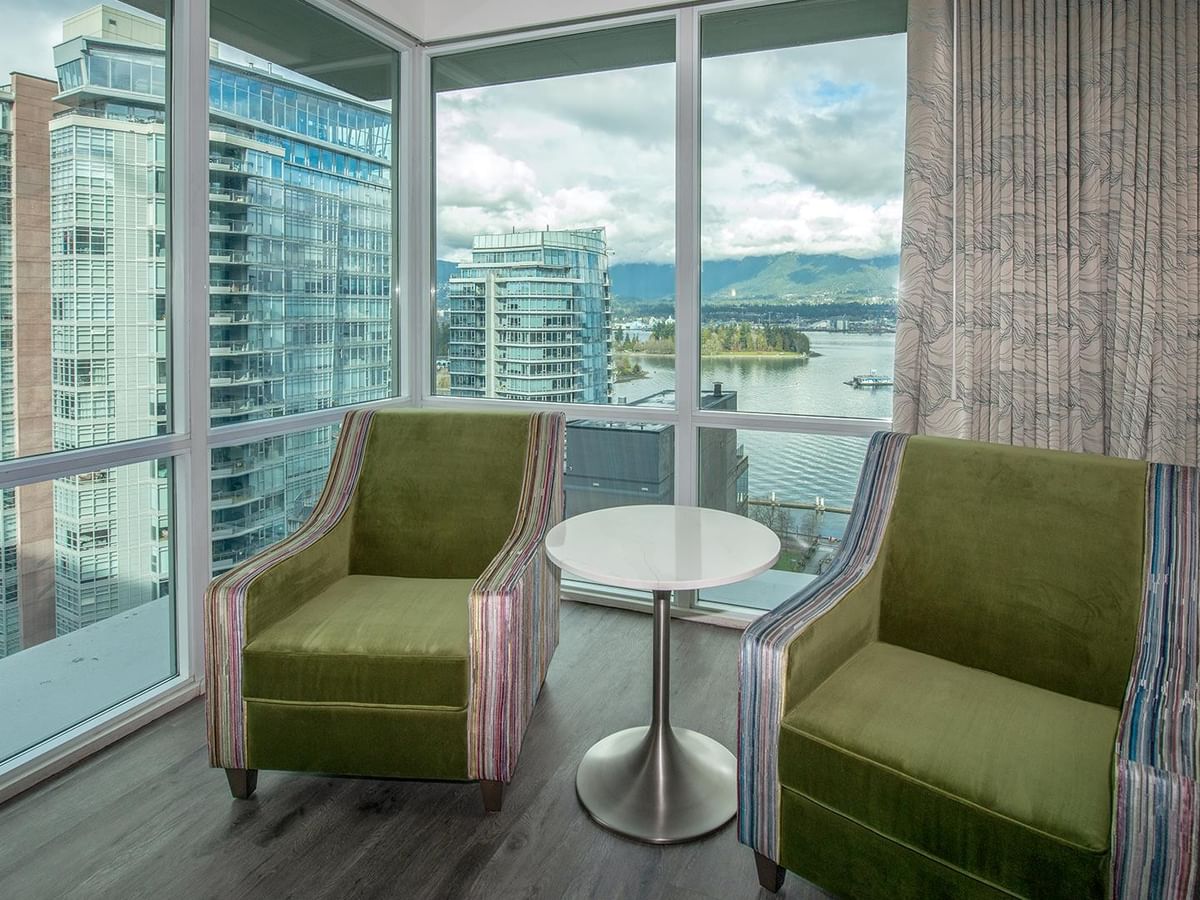Two green chairs, a round table, and a city view from the window.
