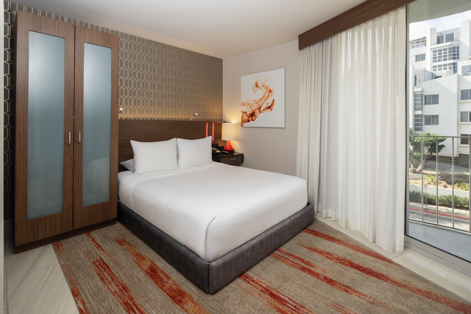 A hotel room with a large bed, wardrobe, nightstand, and artwork