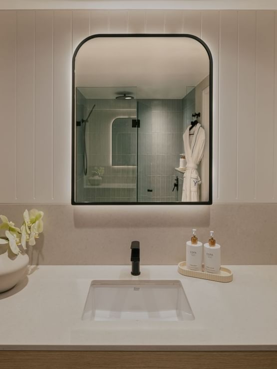 Ocean View Terrace King at Elysium Manly, featuring a sink by lotion bottles under a lit mirror, next to a vase of orchids