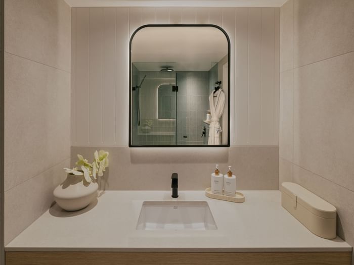 Ocean View King – Elysium Floor at Elysium Manly, featuring a sink with lotion bottles under a lit mirror, next to a vase