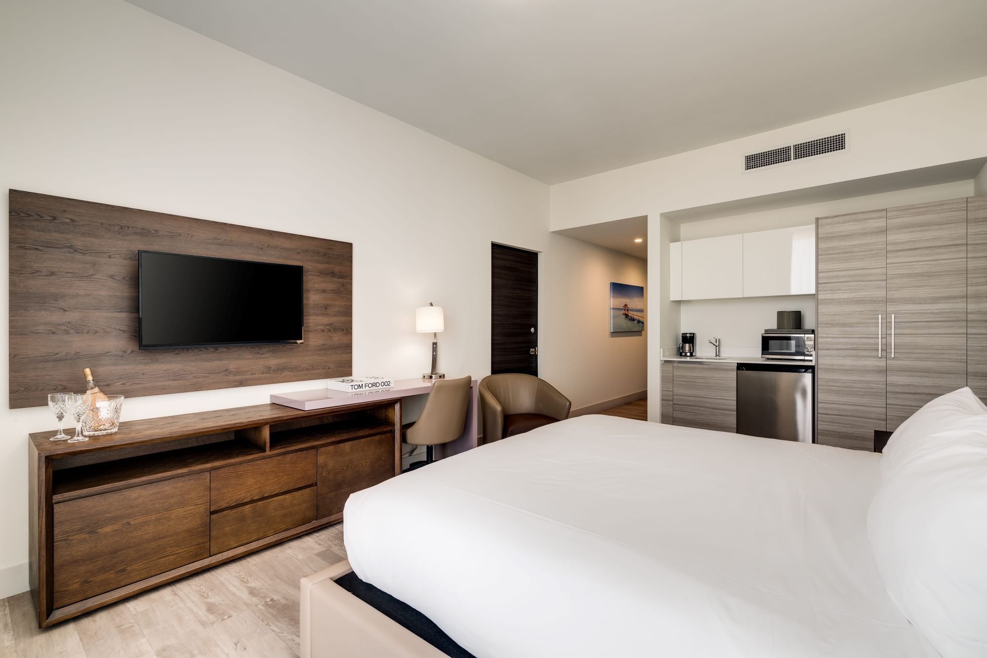A modern hotel room with a large bed, flat screen TV, desk, and kitchenette.