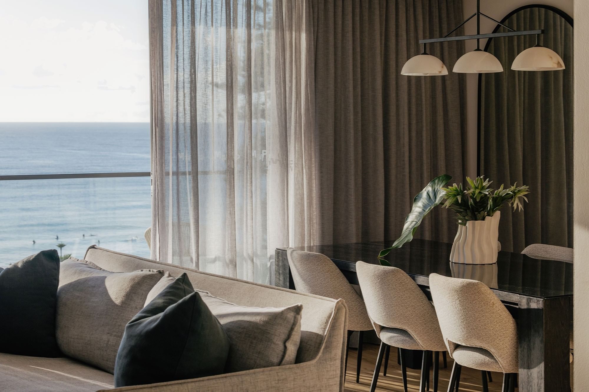 Living room with grey cushions by a dining table under sheer curtains overlooking the ocean at Elysium Hotels