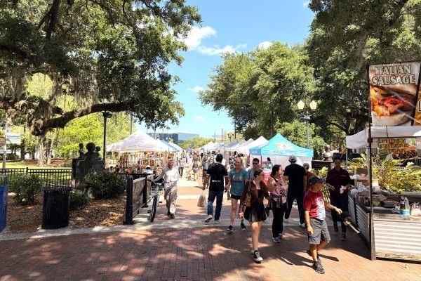 A pathway of canopy tents and a crowd of people beneath large lush trees at Fall Fiesta in the Park in Orlando.