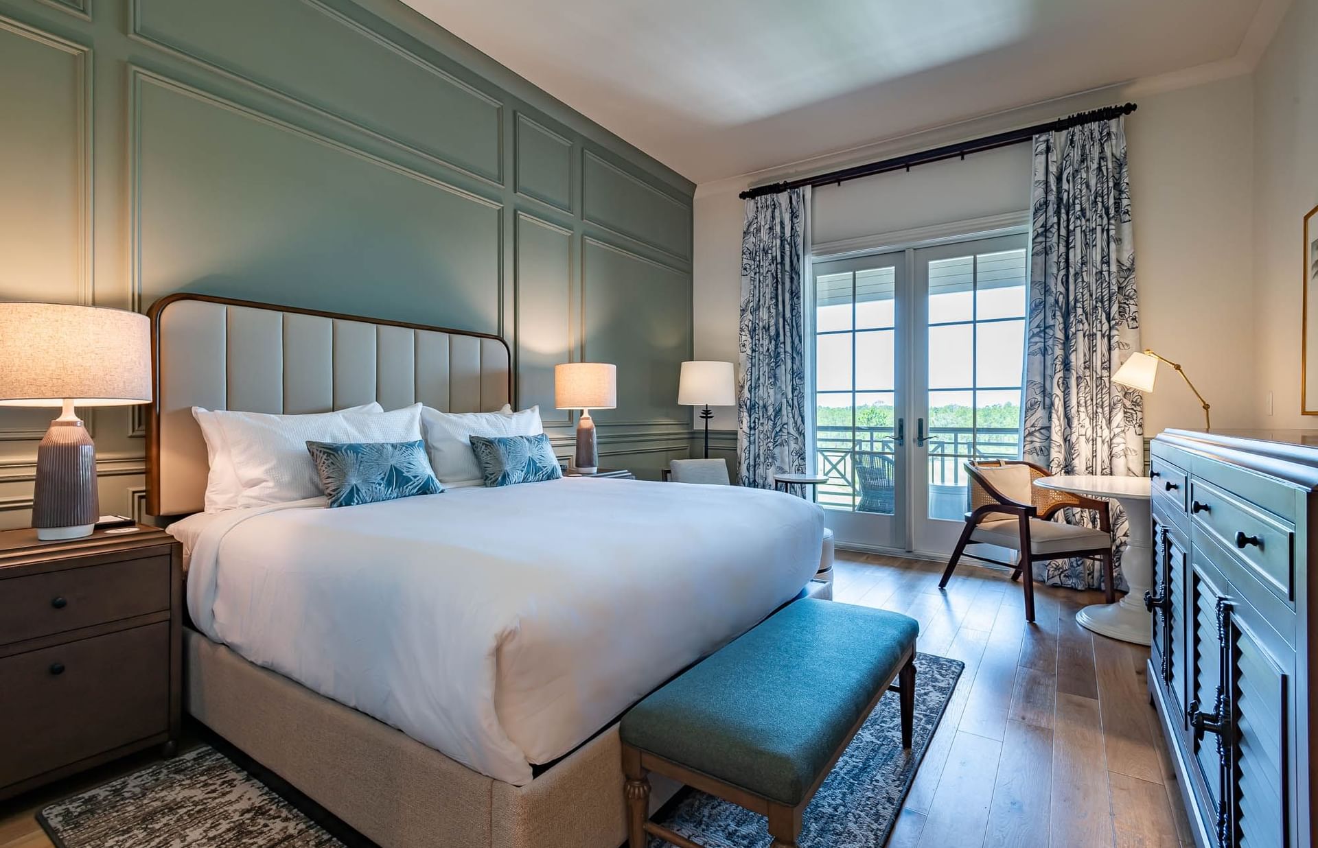 A luxurious bedroom with a large bed, two lamps, a dressing table, and a balcony overlooking a forest.