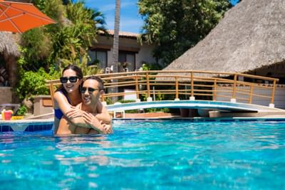 Couple wearing sunglasses embracing in pool with thatched roof huts and wooden bridge in background.