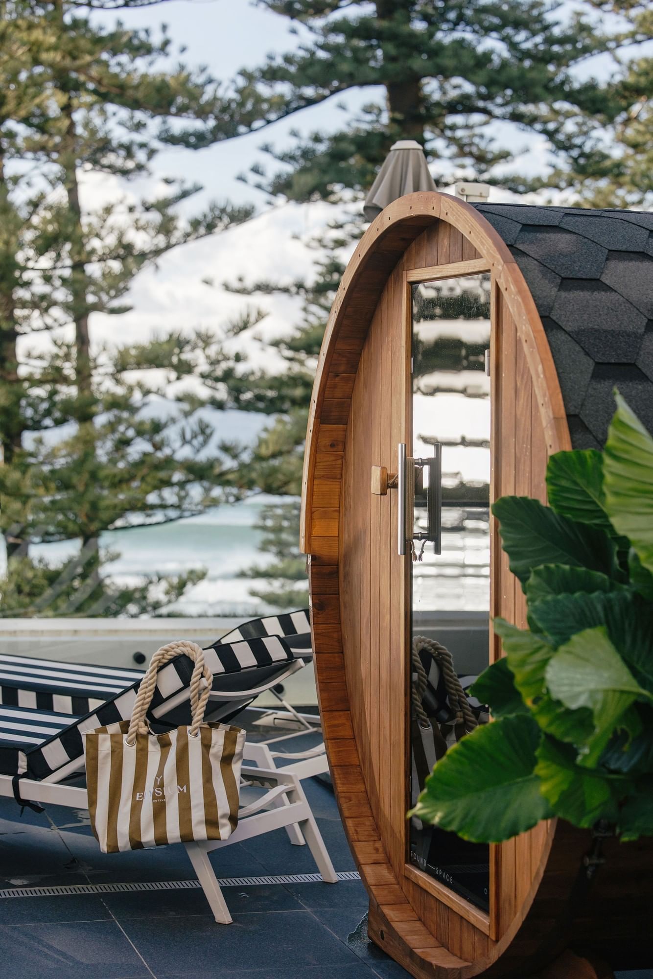 Luxury deck at Elysium Hotels featuring a wooden barrel sauna with striped lounger chair and tote bag near ocean view