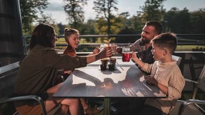 A family of four with two kids is seated at a table outside, toasting with drinks.