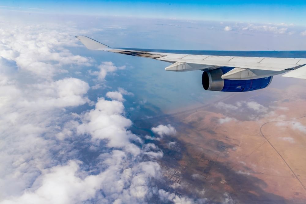 Looking out over an airplane wing flying over blue sky, clouds, and brown ground far below. 