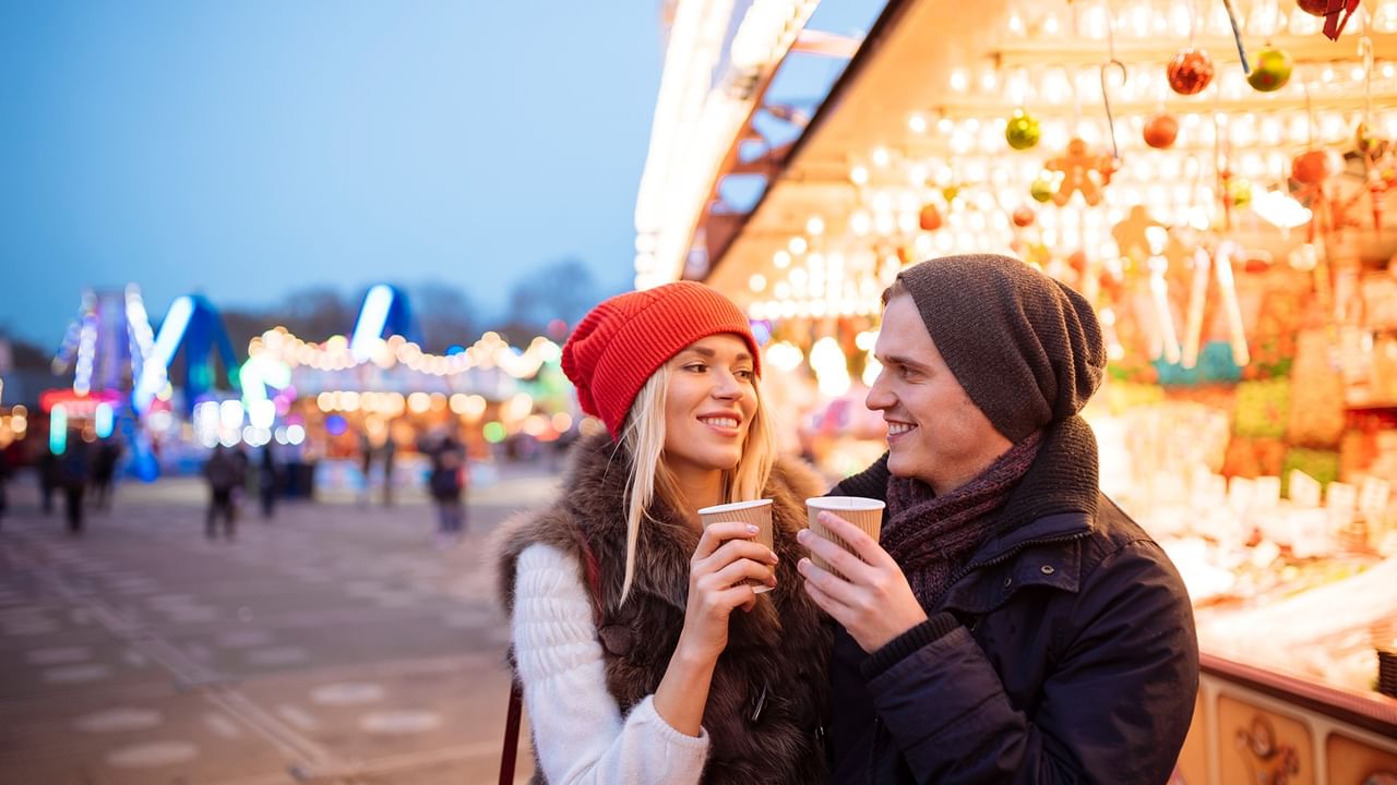 Couple enjoying warm beverages at an illuminated festive market with colorful lights and holiday decorations at dusk.