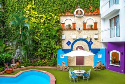 Patio colorido con piscina, muro decorativo con arcos y vegetación tropical en Los Arcos Suites