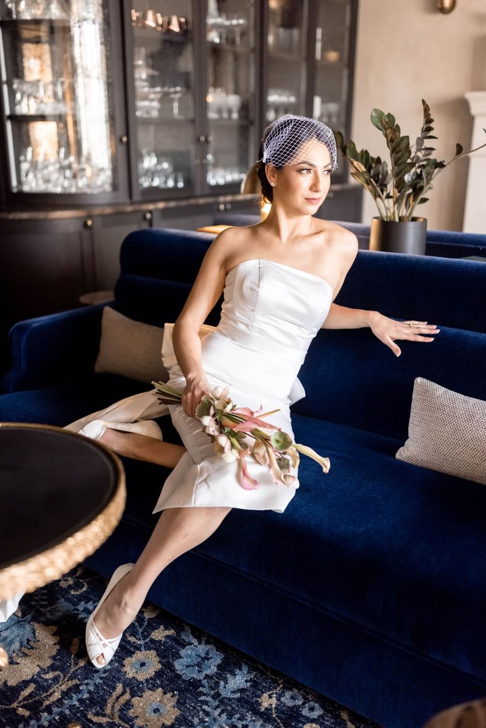Beautiful bride in a white dress and veil, holding a bouquet while seated on a blue sofa at El Prado Hotel