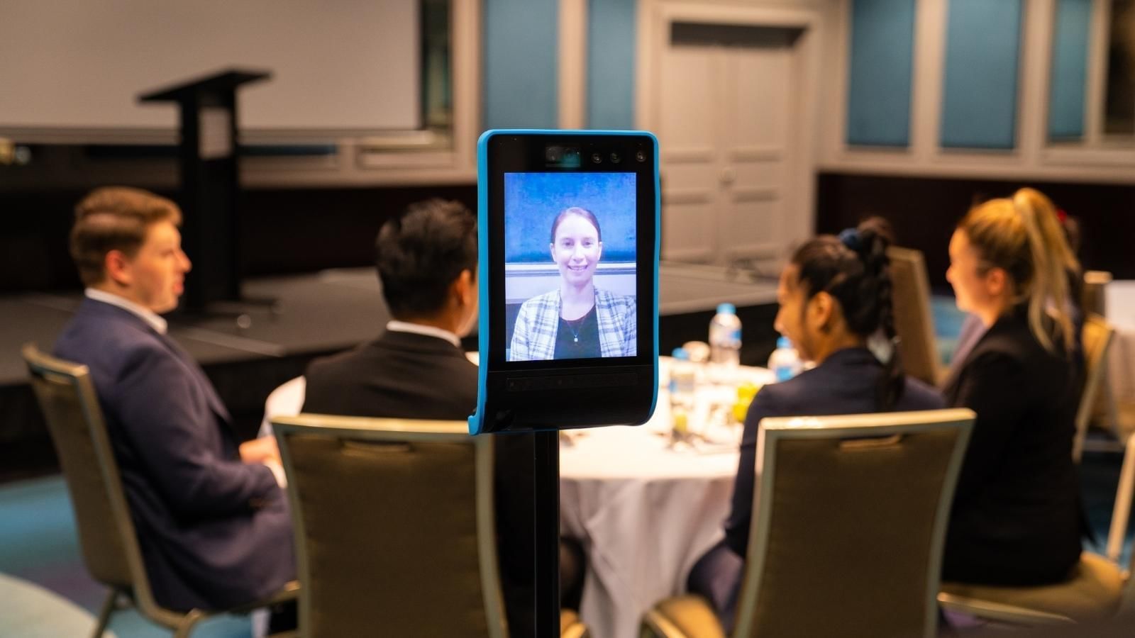 People sitting around a table and a lady joined via remote camera for the meeting at Pullman King George Square