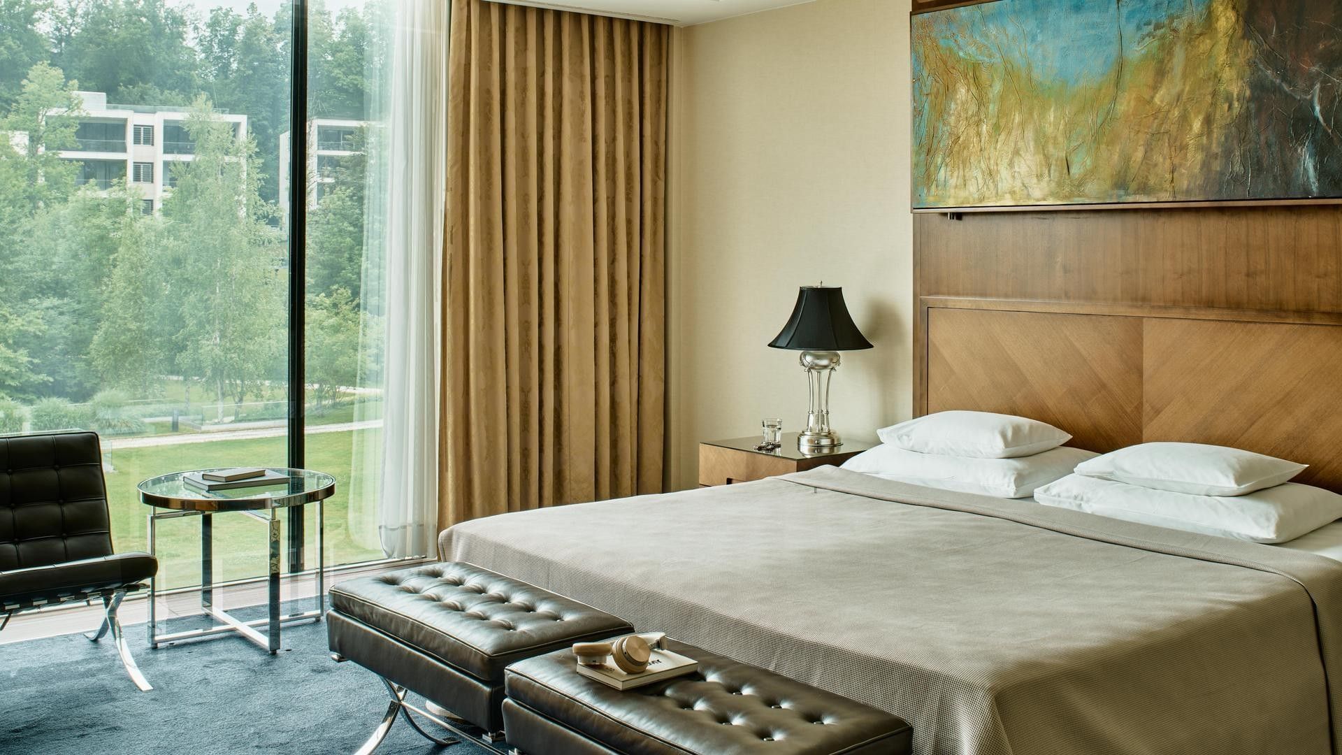 Elegantes Hotelzimmer mit großem Bett, Sesseln, Tisch und großem Fenster mit Blick auf Bäume.