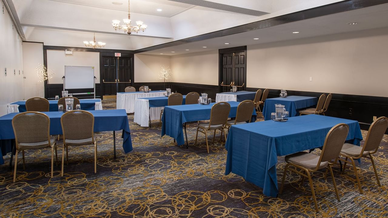 Conference room with blue tablecloths, chairs, carpeted floor, chandeliers, and a whiteboard.