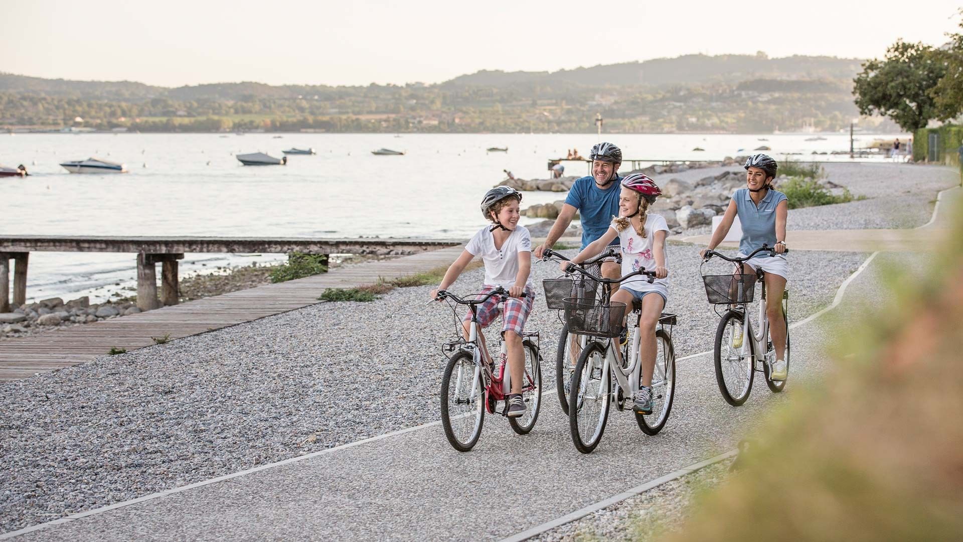 Family cycling by the lake with boats in background at Falkensteiner Resort Lake Garda
