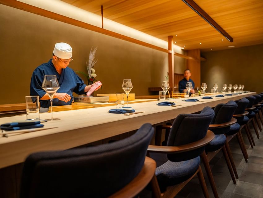 Chef prepares food at a long sushi bar with wine glasses and napkins on the counter.
