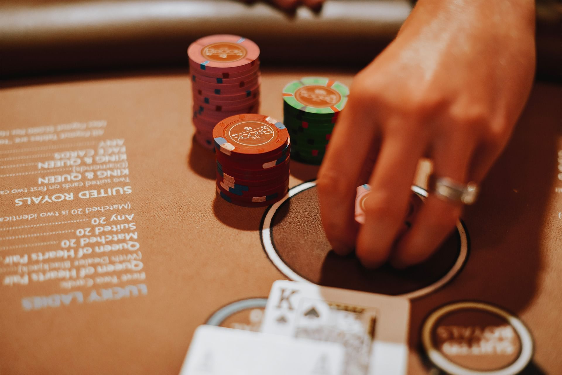 Hand moving poker chips by stacked red, green, and pink chips with playing cards on a gaming table at The Brook