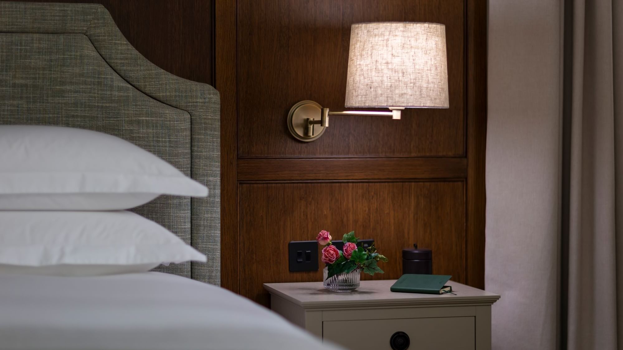 Bed, with a wall-mounted lamp, flower vase & notebook on the nightstand in Twin Garden Suite at Dunluce Lodge